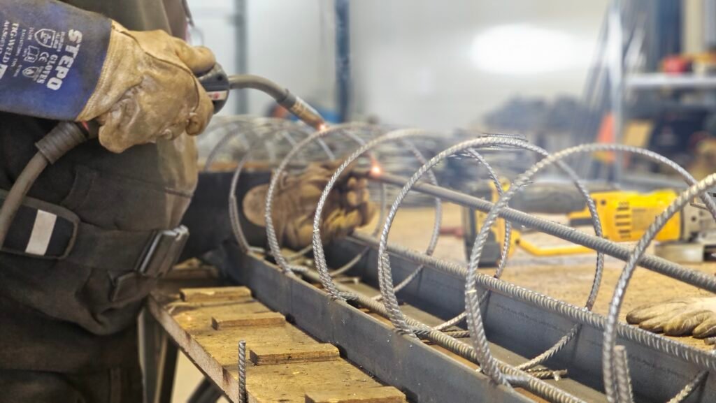 Darbuotojas suvirina metalinę armatūros spiralę dirbtuvėse
